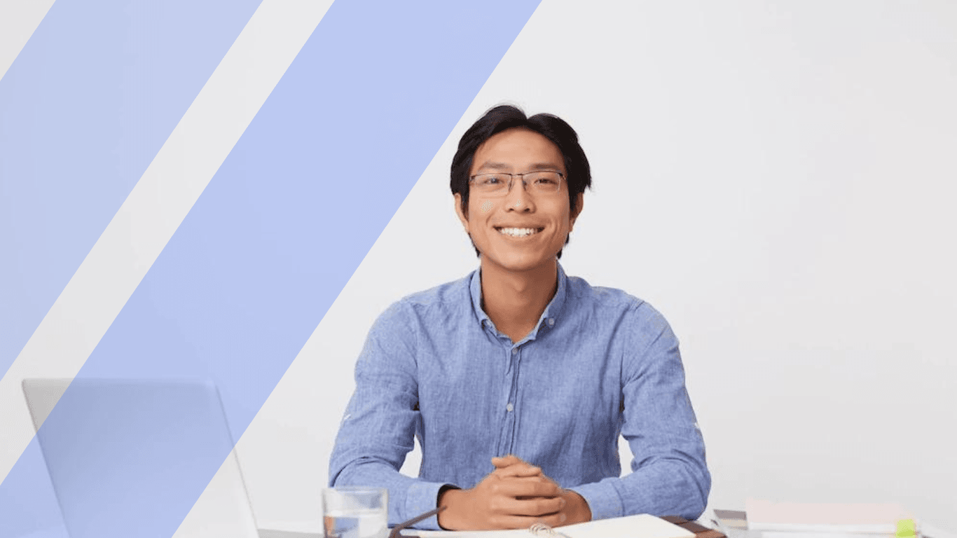 asian young businessman in glasses in blue shirt sitting at the table with laptop over white wall